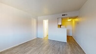 This single-floor unit offers a straightforward, functional layout with consistent light wood-look vinyl flooring running throughout the space. The living area is open and airy, featuring white walls, smooth ceilings with a ceiling vent for climate control, and a window that brings in natural light while providing a view to the exterior. The space connects seamlessly to the kitchen area via a pass-through counter, creating a sense of openness between the two zones.

The galley-style kitchen is efficiently laid out along a single wall, equipped with white cabinetry, a white electric coil-top range with oven, a refrigerator, and a sink set into a speckled countertop. Upper cabinets provide overhead storage, and overhead lighting illuminates the workspace. The warm-toned flooring in the kitchen contrasts slightly with the cooler tones in the living area.

A separate bedroom offers a quiet retreat, with the same consistent flooring, white walls, and access to the hallway. The bathroom is clean and well-appointed, featuring a white vanity with a speckled countertop, chrome hardware, under-sink cabinet storage, a wall-mounted mirror, and a vanity light fixture above. The overall ambiance of the unit is clean, neutral, and move-in ready.