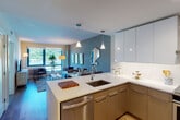 This single-floor residence is designed with a contemporary aesthetic, combining functionality and style throughout its well-planned layout. The open-concept kitchen flows seamlessly into the living and dining areas, creating a cohesive, social space. The kitchen is outfitted with sleek flat-front cabinetry in a two-tone palette of warm taupe and crisp white, stainless steel appliances including a range with hood ventilation and built-in microwave, a white quartz peninsula with an undermount sink, and open shelving with a striking blue interior accent. Pendant lighting above the peninsula adds a refined touch.

The living area is anchored by large windows that frame verdant outdoor views, flooding the space with natural light. Warm hardwood-style flooring runs throughout, complementing the muted blue and sage green accent walls. The primary bedroom features a generous floor-to-ceiling window with expansive views, hardwood flooring, and a dedicated closet. A second bedroom includes closet access and shares the space's calm, neutral color palette. The bathroom is cleanly finished with large-format textured tile in the tub/shower surround and chrome fixtures, maintaining the residence's modern sensibility throughout.