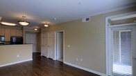 This single-floor space showcases a clean and functional layout highlighted by rich, dark hardwood flooring that runs throughout the visible areas. The walls are painted in a warm beige tone, creating a neutral and inviting atmosphere. Crown molding lines the ceiling perimeter, adding a subtle architectural detail that elevates the overall finish of the space.

The hallway corridor is a notable feature, lined with multiple white six-panel doors that provide access to various rooms. The crisp white trim and door frames contrast neatly against the warm wall color, contributing to a polished, cohesive aesthetic. Ceiling-mounted light fixtures with brushed metal accents provide ambient illumination along the corridor.

HVAC vents are positioned both on the ceiling and walls, ensuring efficient climate control throughout. Electrical outlets are conveniently placed along the walls, reflecting practical, move-in-ready functionality. The open sightlines from the main living area toward the hallway and an adjacent kitchen space suggest a logical, well-organized floor plan. The combination of warm tones, dark flooring, and white accents gives the space a balanced, traditional character.