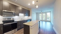 This well-appointed single-floor residence combines modern design with functional living. The kitchen serves as a central highlight, featuring dark espresso cabinetry extending to the ceiling, a classic white subway tile backsplash, granite countertops, and a full suite of stainless steel appliances including a refrigerator, range, microwave, and dishwasher. A granite-topped island with pendant lighting provides additional prep space and casual dining. Warm hardwood-style flooring runs throughout the kitchen and living areas, lending a cohesive and polished aesthetic. The open-concept living area is bright and airy, anchored by a ceiling fan and floor-to-ceiling glass doors that open to a private balcony with green treetop views, filling the space with natural light. The bedrooms are finished with plush carpeting and ceiling fans, offering a comfortable retreat. The primary bedroom includes dual closets with built-in shelving for ample storage. The bathroom features a full soaking tub with white tile surround and clean, crisp finishes. Throughout the residence, neutral tones and thoughtful finishes create a contemporary and welcoming atmosphere suited for modern living.