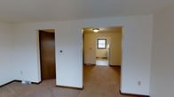 This three-floor space presents a straightforward, functional layout with clean white walls and warm brown trim throughout. The main level, visible in the image, features wall-to-wall carpeting in a neutral beige tone, providing a comfortable underfoot feel across the open living area. A hallway leads into an adjacent room with hard-surface flooring, likely a kitchen or utility area, distinguished by a small window that brings in natural light. A flush-mount ceiling fixture provides overhead illumination in this secondary space. The brown-trimmed doorways and baseboards offer a consistent design detail that grounds the otherwise light, neutral palette. A thermostat is mounted on one of the interior walls, indicating climate control accessibility on this level. A solid wood door with a classic brown finish is visible, adding a traditional touch to the entry or utility access point. The open passage between rooms creates a connected feel while maintaining defined functional zones. Spanning three floors in total, this space offers meaningful vertical separation between living, sleeping, and utility areas, providing a well-organized and versatile layout suitable for a variety of uses.