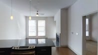 This single-floor space showcases a contemporary open-plan layout that seamlessly connects the kitchen and living areas. The kitchen is equipped with dark cabinetry topped with polished black granite countertops, providing a sleek contrast against the neutral white walls. A stainless steel oven and range are integrated into the kitchen island, offering a functional and stylish cooking area.

The living space beyond is bright and airy, benefiting from two large windows dressed with white blinds that allow generous natural light to fill the room. A ceiling fan with a light fixture is centrally mounted, complementing a pendant light that hangs near the kitchen area, together providing layered lighting options throughout.

A built-in fireplace serves as a focal point on one wall of the living area, adding both architectural interest and a cozy ambiance to the space. Warm-toned hardwood flooring runs throughout, tying the open-plan areas together cohesively. The clean lines, minimal detailing, and neutral color palette give the space a modern, understated character suited to a variety of interior styles.