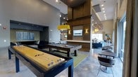 This single-floor amenity lounge is designed as a sophisticated entertainment and social hub, combining modern aesthetics with a variety of recreational offerings. The space commands attention with its soaring ceilings and an open-concept layout that seamlessly connects multiple activity zones.

At the heart of the room, a full-size pool table and a shuffleboard table serve as focal points for recreation, positioned beneath a striking yellow pendant light that adds a bold pop of color against the otherwise neutral palette of grays and rich wood tones. A large wall-mounted flat-screen television is anchored to a floor-to-ceiling wood-paneled feature column, flanked by accent wall sconces that cast warm, inviting light.

The polished concrete flooring enhances the industrial-chic character of the space, while recessed ceiling lighting and decorative pendant fixtures throughout maintain a bright, welcoming atmosphere. To one side, a lounge area with plush dark sofas provides a relaxed retreat, while a dining area with tables and chairs offers additional functionality. Large windows at the far end draw in natural light, contributing to the airy, energetic ambiance of this well-appointed communal space.