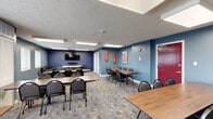 Located at 10129 S Lake Blvd in Parma, Ohio, this single-floor community space offers a well-appointed and multi-functional layout designed for gatherings and social activities. The interior is finished with cool slate-blue and navy accent walls, patterned carpet flooring, and flush-panel ceiling lights that create a clean, modern ambiance throughout.

The combined dining and living area serves as the heart of the space. The living zone features dark leather sofas and chairs arranged around a wall-mounted flat-screen television set against a striking navy geometric accent wall, providing a comfortable lounge setting. Adjacent to this is the dining and game area, which accommodates multiple rectangular tables with seating for groups of varying sizes, alongside a dedicated poker table — making it equally suited for meetings, dining, or recreational use. Windows fitted with white blinds line the walls, allowing natural light to filter in and offering views of the surrounding neighborhood.

The kitchen is a functional galley-style space outfitted with marble-look laminate countertops, ample dark cabinetry, wood-look vinyl plank flooring, a gas range, a full-size refrigerator, and a sink — providing everything needed to support catered events or casual gatherings. Decorative botanical prints add a welcoming touch to the kitchen walls.