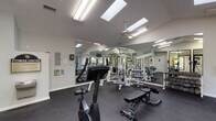Located at 1905 Natchez Trace in Greensboro, NC, this single-story fitness center is designed to support a comprehensive workout experience. The facility is organized into distinct exercise zones, each thoughtfully equipped for different training needs.

The strength training area features a Quantum multi-station cable machine, adjustable benches, and a full dumbbell rack, all set against floor-to-ceiling mirrors that visually expand the space and assist with form. Dark rubber flooring throughout provides cushioning and durability underfoot.

The cardio section is positioned along a wall of large, black-framed windows offering views of lush, tree-lined greenery, creating an inviting environment during workouts. This area houses multiple treadmills and elliptical trainers, as well as upright and recumbent stationary bikes. A wall-mounted television is positioned for convenient viewing.

A dedicated activity room, separated by glass-panel walls and doors, provides space for stretching and functional fitness, stocked with stability balls and accessories. The facility also includes a water fountain for convenience.

Overhead, multiple skylights and vaulted ceilings flood the main workout area with natural light, complemented by ceiling fans and recessed fluorescent lighting. The bright, airy atmosphere and well-rounded equipment selection make this fitness center a practical and motivating space.