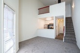 Located in Walnut Creek, CA, this two-story, one-bedroom, one-bathroom home offers a smart and efficient layout spread across two floors. The main level features a soaring vaulted ceiling in the living and dining area, creating an airy, open atmosphere that feels larger than its footprint suggests. Large windows with slatted blinds line the living room walls, inviting abundant natural light and offering views of the surrounding neighborhood. A wall-unit air conditioner and an exterior door provide both comfort and convenient access. The open-concept kitchen is positioned on a raised platform visible from the living area, outfitted with white cabinetry, stainless steel appliances including a refrigerator and microwave, and a breakfast bar-style counter. A staircase with a distinctive red railing leads from the main level to the upper floor, where the private bedroom is situated. The bedroom features plush carpeting and a large window with blinds that filters in soft, natural light. Three closets throughout the home provide generous storage. The full bathroom is finished with wood-look tile flooring, a combined tub and shower with sliding glass doors, a toilet, and a single-sink vanity with a light countertop. The overall aesthetic is clean, bright, and neutral throughout.
