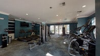 This fitness center in Houston, TX (77027) is a single-floor facility designed to support a comprehensive workout experience. The interior is defined by bold teal-painted walls accented with white crown molding, recessed lighting, and patterned carpet flooring that lends a polished, residential feel to the space.

The cardio zone features a row of treadmills positioned along one wall beneath a wall-mounted flat-screen television, flanked by built-in shelving and cabinetry. Elliptical machines and a stair climber are also available, offering a variety of low-impact cardiovascular options. The strength training area includes a cable crossover machine, a power rack with a flat bench and weight plates, and a multi-use functional trainer, providing options for both machine-based and free-weight workouts. A full dumbbell rack lines the mirrored wall, with a large mirror panel reflecting the space and enhancing the sense of openness.

Natural light enters through multiple windows and glass-paneled doors that open toward an outdoor pool area and courtyard. This connectivity to the outdoors contributes to a bright, airy atmosphere throughout the facility. The overall design balances functionality with aesthetic cohesion, creating an inviting environment suited for a range of fitness routines.