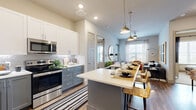 This 2-bedroom, 3-bathroom single-floor residence offers a smart and stylish layout designed for comfortable everyday living. The open-concept kitchen and living area serves as the heart of the home, where a sleek kitchen island with seating anchors the space. The kitchen features two-tone cabinetry — white uppers paired with slate-blue lowers — complemented by a mosaic tile backsplash, stainless steel appliances, and a gooseneck faucet. Pendant lighting adds a refined touch above the island. The adjacent living room is bright and welcoming, with large windows that invite natural light, a ceiling fan, and warm wood-look flooring that extends throughout the main areas. The primary bedroom offers a comfortable retreat with a large window overlooking outdoor views and direct access to a well-appointed en-suite bathroom. The primary bath includes a single vanity with white countertops, dark cabinetry, a walk-in glass shower with tile surround, and a connected walk-in closet. The second bedroom is equally well-sized and cozy. A second full bathroom features a soaking tub and matching vanity finishes. Two dedicated closets and a hallway complete the efficient and functional floor plan.