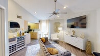 Located at 1855 Craigshire Rd in Maryland Heights, Missouri, this one-bedroom, one-bathroom residence offers a thoughtfully designed layout that maximizes every square foot. The open-concept living area immediately impresses with vaulted ceilings and a ceiling fan, creating an airy, spacious feel. Wood-look flooring runs throughout, anchoring the warm, contemporary aesthetic. A tufted gray sectional sofa, white dresser with gold hardware, and a wall-mounted flat-screen TV furnish the living room, while gold-accented décor and a geometric wall art piece add a curated, modern touch. Natural light filters in through curtained windows, brightening the space. The galley-style kitchen features warm wood cabinetry, white countertops, black appliances, and ample storage on both sides, opening toward a breakfast bar area with seating. The bedroom is a serene retreat, highlighted by a skylight that draws in natural light, complemented by soft carpet flooring, elegant wall sconces, and direct access to the bathroom. The bathroom offers a clean, modern design with a wood-toned vanity, white countertop, large mirror, and a separate tub-shower combination with tile flooring. Every room reflects a cohesive, contemporary design sensibility with gold accents and neutral tones throughout.