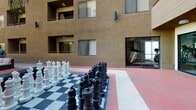 Located at 840 17th St in San Diego's vibrant 92101 corridor, this outdoor amenity courtyard offers a well-appointed communal space designed for both recreation and relaxation. The courtyard's most distinctive feature is a large-format chess set laid out on a bold black-and-white checkered surface, providing an engaging focal point for residents and visitors alike. The surrounding area is finished with a distinctive red-toned court surface that adds a lively visual contrast to the warm stucco exterior of the multi-story building above.

A comfortable lounge area is situated toward one end of the courtyard, furnished with generous sectional seating and shaded by a pergola canopy, making it an ideal spot to unwind under the Southern California sun. Tropical palms and potted greenery contribute a relaxed, resort-like atmosphere to the space.

Direct access to a fully equipped fitness center is available via glass-paneled doors opening directly onto the courtyard. Through the windows, cardio equipment including treadmills and other machines is clearly visible, offering convenience for active residents. The building's textured stucco façade, framed windows, and tiered balconies frame the courtyard on multiple sides, creating an enclosed yet open-air environment that balances privacy with an abundance of natural light.