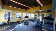 This fitness center offers a focused and functional workout space designed to accommodate a variety of cardio and exercise needs. The room is outfitted with key equipment including a treadmill, elliptical trainer, and stationary bike, providing users with multiple options for cardiovascular training. Large windows along one wall allow natural light to fill the space, creating a bright and motivating atmosphere while offering views of the surrounding exterior. A full-length mirrored wall runs along one side of the room, visually expanding the space and allowing users to monitor their form during workouts. The flooring consists of durable dark rubber matting, ideal for high-traffic fitness use and providing cushioning underfoot. The ceiling features an open industrial design with exposed black structural beams and bold red accent lighting tracks, lending the space a modern, energetic aesthetic. The walls are painted a warm yellow, contributing to the room's vibrant and upbeat ambiance. Overall, this fitness center combines practical equipment, smart design choices, and ample natural light to create an inviting and efficient space for physical activity.