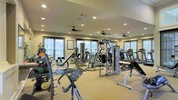 This fitness center offers a thoughtfully designed workout environment spread across a single level. The space is outfitted with a wide variety of exercise equipment, including treadmills, elliptical machines, cable weight systems, and free weights such as kettlebells and medicine balls, catering to a full spectrum of fitness needs. A dedicated bench press station and multi-use cable machine anchor the strength training area, while cardio machines are neatly arranged to maximize usable floor space. The room is finished with a neutral-toned carpet that adds warmth and comfort underfoot. Recessed ceiling lights provide consistent, even illumination throughout, complemented by multiple ceiling fans that ensure comfortable air circulation during workouts. Large windows and glass-paneled doors line multiple walls, flooding the interior with natural light and offering views of the surrounding outdoor greenery. The warm, light-colored walls contribute to an open and inviting atmosphere. Motivational artwork is displayed on the walls, adding a polished finishing touch. Overall, the space strikes a balance between functionality and comfort, making it a well-rounded facility suitable for a wide range of fitness activities.