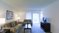 This single-floor space centers around a well-appointed living room designed for comfort and functionality. The living area is finished with wall-to-wall carpeting in a neutral tone, contributing to a warm and quiet atmosphere throughout. A large sliding glass door dominates one wall, fitted with vertical blinds that allow for adjustable natural light control while providing direct access to an outdoor area. The generous amount of natural light that filters through gives the room an open, airy feel.

The living room is arranged to maximize the available space, accommodating a full sofa and a loveseat positioned to face a dark-finished media console supporting a flat-screen television. A rectangular dark wood coffee table anchors the seating area, complemented by a coordinating end table with a lamp that adds warm ambient lighting. A small potted plant on the coffee table introduces a subtle natural accent to the otherwise neutral palette.

The walls are painted a clean white, enhancing the brightness of the room and providing a versatile backdrop. The overall layout is practical and straightforward, with a clear sense of openness that makes the living space feel inviting and easy to navigate.