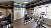 This fitness center offers a comprehensive workout environment spread across a single floor, thoughtfully laid out to accommodate a variety of exercise routines. The space is immediately recognizable by its bold, horizontal striped walls in navy blue, gold, tan, and white — a vibrant design choice that energizes the room and gives it a distinct character.

The flooring throughout is a durable, speckled rubber material well-suited for high-traffic fitness use, providing both comfort and traction. Overhead, pendant-style and flush-mount ceiling lights provide consistent, even illumination across the space, complemented by natural light filtering through windows fitted with blinds.

The cardio zone features multiple elliptical trainers and upright stationary bikes, all equipped with individual display consoles. Strength training is well-represented with a multi-function cable and weight machine, a flat workout bench, and a universal strength station. A large mirror along one wall enhances the sense of space and allows users to monitor their form during workouts.

Practical amenities include a dual wall-mounted water fountain for convenient hydration, a posted fitness area rules sign, and a waste receptacle. A separate anteroom is accessible via an interior door, and clearly marked emergency exits ensure safety. The layout is open and organized, making the fitness center both functional and welcoming.
