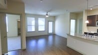 This single-story space presents an open-concept living area defined by rich, dark hardwood-style flooring that runs throughout the main space. The room benefits from abundant natural light, courtesy of multiple windows fitted with horizontal blinds, as well as a glass-paneled door that leads to an exterior access point, further brightening the interior. A ceiling fan with an integrated light fixture is centrally positioned overhead, providing both air circulation and supplemental lighting against the smooth, neutral-toned walls and white ceiling.

The layout flows seamlessly toward a kitchen area, partially visible at the right side of the space. A countertop with granite-style detailing and light cabinetry beneath serves as a transitional element between the living and kitchen zones. A mirrored surface adjacent to the counter adds visual depth to the area. The overall color palette is kept light and neutral, with creamy walls complementing the warm tones of the flooring. The open floor plan, combined with the generous natural light and clean finishes, creates a bright and functional living environment suitable for a variety of furniture arrangements.