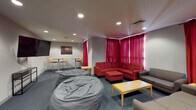 Located at 3009 M E Valentine Dr in Raleigh, NC, this single-floor communal lounge space is designed to foster relaxation, creativity, and social interaction. The room is unified by a cohesive color palette of deep reds and cool grays, anchored by plush carpeted flooring and recessed ceiling lighting throughout.

The seating area is a central highlight, featuring a bold red sectional sofa with a matching ottoman, complemented by gray couches arranged to encourage group gathering. Large, oversized bean bag chairs are scattered throughout, adding a casual and comfortable element to the space. A wooden coffee table and side tables provide functional surfaces for everyday use.

A wall-mounted flat-screen television serves as a media focal point, flanked by framed photographic artwork that adds a curated, artistic touch. Striking red curtains frame multiple windows along one wall, allowing diffused natural light to filter into the space while contributing to its vibrant aesthetic.

One of the most distinctive features is a full-length chalkboard mural wall, covered in elaborate, hand-drawn artwork including abstract figures, flora, and celestial motifs — serving as both a creative canvas and a dynamic visual centerpiece. A high-top table with bar stools offers an additional casual seating zone. The space is accessible via glass-paneled doors, adding a modern, open feel to the overall design.