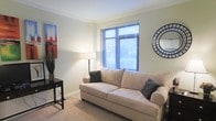 This single-floor residential space offers a comfortable and stylish living environment with thoughtful design details throughout. The living area serves as the centerpiece of the space, showcasing elegant crown molding that adds a refined architectural touch to the room. Neutral, light-toned walls create a warm and inviting atmosphere, complemented by plush carpeted flooring underfoot. A large window fitted with horizontal blinds allows natural light to fill the room while providing privacy, offering a glimpse of lush greenery beyond. The space is furnished with a classic cream-toned sofa accented with decorative pillows, creating a cozy and welcoming seating area. A sleek black side table, a contemporary floor lamp, and a decorative mirror with a sunburst-style frame add character and balance to the room. A bold piece of wall art injects a pop of color, lending personality to the otherwise softly toned interior. The overall ambiance is clean, bright, and well-maintained, reflecting a space that blends comfort with understated elegance. The single-floor layout ensures easy accessibility and a cohesive flow throughout the living environment.