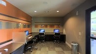 This single-floor business center is designed to provide a functional and visually engaging workspace. The room features three iMac desktop computers arranged along a continuous dark wood desk that spans the perimeter, each station paired with a sleek, adjustable ergonomic office chair, offering comfortable seating for extended use. The space is defined by a striking color palette, combining warm burnt orange accent walls with neutral taupe tones, giving it a contemporary and energetic feel. Horizontal striped wall art with small accent lighting adds a decorative focal point along the back wall, enhancing the room's polished aesthetic. Recessed ceiling lighting provides consistent, even illumination throughout, complementing the light-toned tile flooring laid in a subtle checkerboard pattern. The layout is efficient and well-organized, making it easy for multiple users to work simultaneously without feeling crowded. A small waste receptacle is positioned near the entrance for convenience. Overall, the space balances practical functionality with modern design sensibilities, making it well-suited for productivity tasks in a shared-use setting.