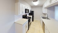 This single-floor unit offers a practical and well-organized layout designed for comfortable everyday living. The open-concept floor plan seamlessly connects the living area to the kitchen, creating a sense of spaciousness throughout the main living space. Light beige ceramic tile flooring runs consistently through the living and kitchen areas, complementing the neutral gray walls and white baseboards that define the interior aesthetic.

The kitchen is well-equipped with white shaker-style cabinetry offering ample storage, white countertops, and a white subway tile backsplash. Black appliances, including an electric range, over-the-range microwave, and refrigerator, provide a crisp contrast to the bright cabinetry. A peninsula with countertop seating separates the kitchen from the living area, adding both functionality and definition to the space.

The living area features a window fitted with horizontal blinds, allowing for natural light and privacy control. A white entry door with direct exterior access is also located in this area.

Toward the rear of the unit, the flooring transitions to wood-look vinyl planking, leading to a dedicated laundry area equipped with a stacked washer and dryer unit alongside built-in shelving. Adjacent to the laundry area is a full bathroom featuring a sink with vanity, toilet, and wall-mounted lighting.