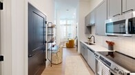 Located at 120 Kindley St in downtown Raleigh, NC, this one-bedroom, one-bathroom residence offers a thoughtfully designed single-floor layout that maximizes every inch of its compact footprint. The galley-style kitchen is a standout feature, outfitted with grey shaker cabinets, white subway tile backsplash, quartz countertops, a matte black faucet, and a full suite of stainless steel appliances including a refrigerator with ice maker, range, dishwasher, and built-in microwave. Light wood flooring runs throughout, lending warmth and continuity to the open flow between the kitchen and living area. The living space benefits from large windows dressed with sheer white curtains, allowing abundant natural light to fill the room. The generously sized primary bedroom features soaring ceilings, tall windows with exterior views, a glass door providing additional light, and direct access to the full bathroom. The bathroom is finished with a floating vanity, quartz countertop, undermount sink, matte black fixtures, and a framed mirror, all complemented by clean white walls. Bold dark interior doors provide a striking design contrast throughout. The overall aesthetic is modern and cohesive, blending contemporary finishes with a bright, airy ambiance.
