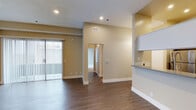This single-floor space showcases a clean, functional layout centered around an open living area. Dark wood-style laminate flooring runs throughout, providing a warm contrast to the neutral beige walls and bright white baseboards. Recessed lighting is installed throughout the ceiling, which features a textured finish, ensuring consistent illumination across the space.

A sliding glass door fitted with vertical blinds lines one wall of the living area, allowing natural light to filter in while offering a connection to the exterior. The ceiling height is notably elevated in the main living zone, contributing to an airy, open feel.

A half-wall peninsula on one side hints at an adjacent kitchen area, where white upper cabinetry and a tiled backsplash are partially visible, suggesting a practical and efficient kitchen design.

A central hallway extends from the living area, leading to additional rooms beyond. The corridor is well-lit and accessible, with a thermostat panel mounted on the wall. The overall layout is straightforward and efficiently organized, making good use of the available floor space while maintaining an uncluttered, move-in-ready appearance.