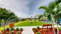 Located at 4003 Oleander Ave in Fort Pierce, Florida, this two-level outdoor amenity complex offers a comprehensive range of recreational facilities designed for active community living. The grounds are thoughtfully landscaped with tropical palms, ornamental grasses, and vibrant flowering plants set against rich red mulch beds, creating a visually appealing Florida aesthetic throughout.

At the heart of the complex sits a generously sized swimming pool with clear turquoise water, enclosed by a secure black aluminum fence and overlooking a natural wooded backdrop. Adjacent to the pool is a dedicated pool house structure with a clean, modern roofline, providing functional support for pool operations.

An outdoor fitness area features a multi-station exercise apparatus installed on a lush artificial turf surface, accompanied by seating benches and interlocking paver pathways that connect the various zones of the property. This area is well-suited for residents seeking open-air workout opportunities.

A dedicated children's playground is situated within its own fenced enclosure, equipped with a multi-level play structure featuring slides, climbing elements, and umbrella-style shade canopies, all set on a safe gravel surface. The perimeter of the complex is secured by consistent black iron fencing, and utility infrastructure is neatly organized on-site. The entire complex benefits from abundant natural Florida sunlight and open skies.