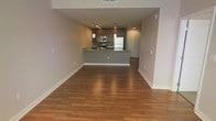 This single-floor residence showcases an open-concept layout that seamlessly connects the living area to the kitchen. The flooring throughout is warm-toned hardwood, adding a sense of cohesion and elegance to the space. Neutral gray and beige walls complement the overall design, creating a clean and modern aesthetic.

The kitchen is a standout feature, equipped with stainless steel appliances including a refrigerator, microwave, and range. Dark espresso-toned cabinetry provides ample storage, while recessed and pendant lighting illuminate the workspace effectively. A raised breakfast bar serves as a natural divider between the kitchen and the living area, offering both a casual dining option and additional counter space. The countertops appear to be a dark stone or laminate finish, complementing the cabinetry.

The open living space is generously sized, providing flexibility for various furniture arrangements. Recessed lighting throughout ensures consistent illumination. A door visible near the kitchen suggests access to a utility or entry area. The overall ambiance is contemporary, functional, and well-designed for comfortable everyday living.