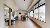 Located at 1410 Deer Lakes Dr in Buffalo, NY, this well-appointed single-floor community space offers a thoughtfully designed layout that balances comfort and functionality. The centerpiece of the space is the expansive living room, distinguished by soaring vaulted ceilings and a large ceiling fan that enhances the open, airy atmosphere. Warm hardwood-style flooring runs throughout, complementing the neutral, contemporary color palette. A stone-surround linear fireplace serves as a striking focal point, topped with nature-inspired artwork that echoes the surrounding environment. Multiple seating arrangements — including a plush sofa, patterned armchairs, and side tables — create inviting areas for gathering and relaxation. A wall-mounted flat-screen television adds a practical entertainment element. Large windows along one wall flood the dining area with natural light and offer views of the outdoors, where additional table-and-chair seating is arranged for communal dining. The kitchen is fully equipped with white shaker cabinetry, ample counter space, black appliances including a refrigerator, range, and over-the-range microwave, and durable tile flooring. A compact, private office provides a dedicated workspace complete with a built-in desk, computer workstation, and greenery accents. The space also includes a comfortable lounge nook near an exterior access door, ideal for a quiet retreat.