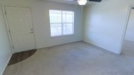 This single-story space presents a straightforward and functional layout highlighted by a well-proportioned main room. The room features neutral, light gray walls that create a calm and versatile backdrop, complemented by wall-to-wall carpeting in a matching neutral tone that adds warmth underfoot. A large multi-pane window along one wall allows ample natural light to fill the space, offering views of an outdoor balcony or patio area beyond, adding a sense of openness to the interior. A ceiling fan is installed overhead, providing both air circulation and ambient lighting. Clean white baseboards trim the perimeter of the room, contributing to the tidy, polished appearance of the space. A door on the left side of the room — featuring a standard handle and a partial view of a hard-surface entry area — provides access to an adjacent area or entry point. The overall ambiance is bright, neutral, and ready for personalization, making it a practical and adaptable living environment. The single-floor layout ensures ease of movement throughout the space.