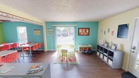 Located at 11201 E Lake Highlands Dr in Dallas, TX, this single-floor educational facility is thoughtfully designed to create an engaging and stimulating environment for young learners. The space is organized into several distinct functional areas, each featuring bold, vibrant wall colors — including lively teal and bright green — that contribute to an energetic and welcoming atmosphere throughout.

The main learning area is anchored by a large sliding glass door that floods the room with natural light, offering views of the surrounding exterior. A child-sized round table and chairs sit atop a colorful area rug, providing an inviting space for early childhood activities, while open shelving units along the walls keep books and learning materials organized and accessible. Educational displays and bulletin boards are mounted throughout, reinforcing a structured yet playful learning environment.

A second classroom features rows of white rectangular tables paired with vibrant red chairs, accompanied by computer workstations along the walls — ideal for group instruction and individual study. A ceiling fan provides comfortable air circulation, and wall-mounted educational maps add to the informative décor.

An administrative area transitions seamlessly from the classrooms, complete with a desk workstation and an adjacent kitchenette equipped with standard appliances and warm wood cabinetry. Durable hardwood-style flooring runs consistently throughout the entire space, tying all areas together cohesively.