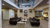 This inviting communal space centers around a striking stacked-stone fireplace wall that rises toward a soaring double-height ceiling, immediately drawing the eye upward and creating a grand sense of openness. A decorative round mirror mounted above the fireplace adds an elegant focal point, while built-in stone shelving with greenery accents frames the feature wall on both sides. The lounge seating area is furnished with deep, dark leather sofas and patterned accent chairs arranged around a central coffee table, offering a comfortable and welcoming atmosphere for relaxation or conversation. An open upper-level mezzanine with a metal railing overlooks the main floor, adding architectural depth and visual interest. Natural and ambient lighting work together to keep the space bright and airy. Beyond the central seating area, additional amenity zones are visible, including what appears to be a game area with a ping pong table and a casual bar or lounge nook with pendant lighting, suggesting this is a multi-functional community gathering space. Motivational signage on the upper walls contributes to an energetic and community-focused ambiance throughout.