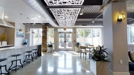 This contemporary lounge is a thoughtfully designed communal space that seamlessly blends industrial and modern aesthetics. The polished concrete floors reflect abundant natural light that streams in through expansive floor-to-ceiling windows, creating a bright and welcoming atmosphere throughout the day. A striking decorative laser-cut ceiling panel serves as a central design focal point, contrasting elegantly against the exposed black ceiling with visible ductwork — a nod to the space's industrial character. A stacked stone accent column adds warmth and texture, grounding the open layout with a natural element. The lounge is furnished with high-top bar seating along one side and casual dining tables with chairs on the other, offering flexible options for socializing or working. Lush potted plants are strategically placed throughout, introducing greenery that softens the clean, minimalist palette of whites and grays. Glass double doors at the far end open directly to an outdoor pool area, making the transition between indoor and outdoor living effortless. This versatile lounge is well-suited for both relaxed gathering and productive use, offering a polished yet comfortable environment with thoughtful architectural details at every turn.