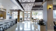 This contemporary lounge is a thoughtfully designed communal space that seamlessly blends industrial and modern aesthetics. The polished concrete floors reflect abundant natural light that streams in through expansive floor-to-ceiling windows, creating a bright and welcoming atmosphere throughout the day. A striking decorative laser-cut ceiling panel serves as a central design focal point, contrasting elegantly against the exposed black ceiling with visible ductwork — a nod to the space's industrial character. A stacked stone accent column adds warmth and texture, grounding the open layout with a natural element. The lounge is furnished with high-top bar seating along one side and casual dining tables with chairs on the other, offering flexible options for socializing or working. Lush potted plants are strategically placed throughout, introducing greenery that softens the clean, minimalist palette of whites and grays. Glass double doors at the far end open directly to an outdoor pool area, making the transition between indoor and outdoor living effortless. This versatile lounge is well-suited for both relaxed gathering and productive use, offering a polished yet comfortable environment with thoughtful architectural details at every turn.