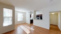 This single-floor space showcases an open-concept layout that seamlessly connects the living area and kitchen, creating a functional and inviting environment. The living area is bathed in natural light streaming through a large window fitted with blinds, casting warm light across the smooth hardwood-style flooring that extends throughout the space. A pendant light fixture hangs above, adding a subtle decorative touch to the dining or living zone.

The kitchen is positioned toward the rear and features rich, dark wood cabinetry that contrasts cleanly against white walls and light-toned countertops. Black appliances, including a refrigerator and microwave, complement the cabinetry's darker tones. A breakfast bar with a pass-through opening connects the kitchen to the main living area, enhancing the open flow and providing additional counter space for casual dining or entertaining. A second window in the kitchen allows additional natural light to filter in. The crisp white walls and ceilings throughout the space contribute to a clean, airy aesthetic, while the warm flooring grounds the overall design with a welcoming, cohesive feel.
