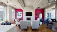 This single-floor community lounge space makes a striking first impression with its bold, magenta accent walls that punctuate an otherwise neutral and sophisticated palette. The main lounge area is anchored by a stunning floor-to-ceiling marble-clad fireplace surround, above which a large flat-screen television is mounted, creating an inviting entertainment focal point. A coffered ceiling with recessed lighting adds architectural depth and elegance to the room. Comfortable light gray upholstered armchairs and a tufted metallic ottoman are arranged on a plush area rug, encouraging relaxed conversation and gathering. Warm hardwood flooring runs throughout the space, complementing the modern yet welcoming aesthetic. Large windows dressed with patterned valances flood the lounge with natural light and offer views of mature trees and the surrounding outdoor landscape. A ceiling fan enhances the airy feel of the sunlit seating area. A dedicated kitchenette features sleek dark cabinetry, white quartz countertops, a stainless steel sink, and a decorative gold-framed mirror, providing practical convenience for events and daily use. Tall indoor plants and curated artwork add organic warmth and personality throughout, making this a well-appointed, design-forward space ideal for community gatherings.