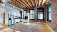 Located at 87 Washington St in Haverhill, MA, this thoughtfully renovated industrial loft seamlessly blends historic character with contemporary design. The open-plan layout is anchored by soaring ceilings showcasing original exposed wooden beams and visible ductwork, underscoring the building's industrial heritage. Original red brick walls provide a warm, textured backdrop throughout, complemented by wide-plank light hardwood flooring that adds warmth and continuity across the space. Tall, arched black-framed windows flood the interior with generous natural light while offering views of the surrounding streetscape. The kitchen is sleek and fully equipped, featuring high-gloss white cabinetry, dark countertops, a hexagonal tile backsplash, and stainless steel appliances including a refrigerator, wall oven, microwave, and dishwasher. The open living area is expansive and flexible, ideal for a variety of uses. A private room with its own black-framed window and white walls provides a quieter, enclosed space. The bathroom is clean and accessible, finished with classic black-and-white mosaic tile flooring, a wall-mounted sink, and matte black fixtures. This space offers a compelling combination of raw architectural detail and modern comfort.