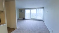 This single-floor space features a generously sized main living area finished with neutral-toned wall-to-wall carpeting that adds warmth underfoot. The room's most prominent feature is a large sliding glass door that spans much of the exterior wall, fitted with vertical blinds and opening to a balcony with outdoor views of surrounding greenery. This expansive window arrangement allows substantial natural light to fill the interior, creating a bright and airy atmosphere throughout the day. The walls are painted in a clean, neutral palette, contributing to the open feel of the space. Climate control is addressed through a wall-mounted air conditioning unit positioned on one side of the room, while baseboard heaters run along the base of the exterior wall, ensuring year-round comfort. A partial wall or entry column subtly defines the transition between the living area and an adjoining section of the space, adding a degree of architectural structure. The overall design is straightforward and functional, offering a clean, move-in-ready environment with practical amenities and a connection to the outdoors through its prominent glazed exterior wall.
