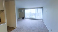 This single-floor space features a generously sized main living area finished with neutral-toned wall-to-wall carpeting that adds warmth underfoot. The room's most prominent feature is a large sliding glass door that spans much of the exterior wall, fitted with vertical blinds and opening to a balcony with outdoor views of surrounding greenery. This expansive window arrangement allows substantial natural light to fill the interior, creating a bright and airy atmosphere throughout the day. The walls are painted in a clean, neutral palette, contributing to the open feel of the space. Climate control is addressed through a wall-mounted air conditioning unit positioned on one side of the room, while baseboard heaters run along the base of the exterior wall, ensuring year-round comfort. A partial wall or entry column subtly defines the transition between the living area and an adjoining section of the space, adding a degree of architectural structure. The overall design is straightforward and functional, offering a clean, move-in-ready environment with practical amenities and a connection to the outdoors through its prominent glazed exterior wall.