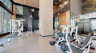 Located in the heart of Midtown Atlanta at 1025 Spring St NW, this contemporary fitness center offers a generously proportioned workout space designed to inspire and perform. The facility is defined by its dramatic double-height ceilings, which create an open, airy atmosphere that sets it apart from typical gym environments. Floor-to-ceiling glass walls line much of the perimeter, flooding the interior with abundant natural light and providing unobstructed views of the surrounding urban streetscape.

The space is divided into distinct functional zones. The strength training area is comprehensively outfitted with a full rack of dumbbells, multiple adjustable benches, weight plates, and a variety of cable and resistance machines. A dedicated free weights section supports a wide range of compound and isolation exercises. Large mirrors are strategically placed throughout, enhancing both visibility and the sense of spaciousness.

Architecturally, the design blends dark textured wall panels with warm wood-toned accents and an eye-catching artistic ceiling installation featuring an intricate gold wire motif. The polished concrete flooring provides a durable, easy-to-maintain surface suited to high-intensity use. A separate glass-enclosed room is accessible from the main floor, offering additional private workout or wellness space. The overall ambiance is upscale, modern, and energizing.