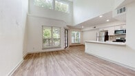 This two-story space is defined by its dramatic double-height ceilings in the main living area, creating an expansive, light-filled atmosphere. A row of clerestory windows positioned at the upper level floods the living space with abundant natural light, complemented by additional windows and a glass-paned door that frames lush, tree-lined outdoor views. Light wood-tone luxury vinyl plank flooring runs throughout the main level, contributing to a clean, contemporary aesthetic.

The open-concept layout seamlessly connects the living area to the kitchen, which features white shaker-style cabinetry, stainless steel appliances including a refrigerator, electric range, microwave, and dishwasher, as well as a white peninsula countertop that serves as both a workspace and informal dining surface.

The upper level transitions to plush carpeting and is equipped with a ceiling fan and recessed lighting. This floor provides access to additional rooms via a paneled interior door, with a white-railed staircase connecting the two levels.

Two bathrooms are present: one featuring a tiled tub-shower combination with subway-style tile and white cabinetry, and another offering a glass-enclosed walk-in shower with matching tile, wood-look flooring, and a white vanity.