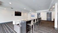 This single-floor communal space is designed with a clean, contemporary aesthetic that prioritizes comfort and social interaction. The layout is anchored by a sleek, dark bar-height counter with seating for multiple guests, making it well-suited for both casual gatherings and social events. A row of modern white and grey high-back bar stools lines the counter, complementing the overall neutral color palette of the room.

A wall-mounted flat-screen television is positioned prominently for easy viewing from multiple seating areas. The lounge seating area features plush grey and white upholstered furniture, providing a relaxed atmosphere for conversation or entertainment. Recessed lighting throughout the ceiling offers consistent, even illumination across the space.

The flooring consists of a bold, multi-toned striped carpet that adds visual interest and a touch of dynamic energy to the room. White walls adorned with a single piece of abstract wall art contribute to the bright and open feel of the space. A textured ceiling adds a subtle architectural detail overhead.

An exit door is visible in the background, indicating this space is part of a larger building complex. Overall, this lounge combines functional design with modern style, creating an inviting environment ideal for community use or hospitality purposes.