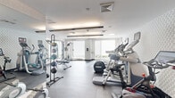 Located at 240 Sidney St in Cambridge, MA, this fitness center offers a thoughtfully designed workout space that balances functionality with a clean, modern aesthetic. The single-floor facility makes efficient use of its footprint, providing a comprehensive range of exercise options for users of all fitness levels.

The cardio area is lined with a row of treadmills, elliptical trainers, and stationary bikes — including Peloton cycles equipped with large interactive screens — all positioned to take advantage of the natural light streaming in through large windows fitted with white horizontal blinds. Patterned accent walls add a subtle visual interest to the space without detracting from its crisp, neutral color palette of white and light grey.

The strength training zone features a multi-cable machine, a weight bench, medicine ball racks, and a rowing machine, catering to a full range of resistance and functional training exercises. Full-length mirrors line one wall, visually expanding the space and allowing users to monitor their form during workouts.

Overhead, recessed and linear lighting fixtures maintain a bright, welcoming atmosphere throughout. The durable dark grey flooring provides both safety and practicality. The space is completed by a key-card access entry door, underscoring its role as a dedicated, secure amenity facility.