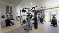This modern fitness center is thoughtfully designed to support a wide range of workout routines across its single-floor layout. The space is divided into distinct functional zones, each tailored to a specific type of training. The strength and cardio area features a Life Fitness Synrgy functional trainer, a TKO heavy punching bag, and a stair climber, all set against floor-to-ceiling mirrors that amplify the sense of space. Rubberized flooring provides durability and comfort underfoot, while recessed lighting and a large ceiling fan ensure the area stays well-lit and well-ventilated. A wall-mounted flat-screen TV adds entertainment during workouts. Adjacent to the main training floor, the spin studio stands out with its light hardwood flooring, expansive floor-to-ceiling windows that flood the room with natural light and outdoor views, and a row of stationary cycling bikes arranged in front of a large display screen. A statement multi-blade ceiling fan anchors the room overhead. A dumbbell rack lines the mirrored wall, offering convenient access to free weights. The overall ambiance is clean, bright, and energizing, making this fitness center a well-rounded amenity suited for users of all fitness levels.