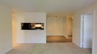 This single-floor residence offers a thoughtfully designed open-concept layout that maximizes the sense of space and flow. The living area is finished with soft wall-to-wall carpeting, creating a comfortable and inviting atmosphere. A defining architectural feature is the built-in pass-through kitchen window, which connects the kitchen to the living space while maintaining a degree of separation. The kitchen is equipped with dark appliances, including a refrigerator and range, complemented by warm wood cabinetry and an under-cabinet range hood that provides focused task lighting. Adjacent to the living area, the space transitions to a hardwood-style flooring, distinguishing a secondary living or dining zone. This area benefits from a track lighting fixture overhead, offering practical illumination. Clean white walls and white trim run throughout the unit, lending a bright and neutral palette that adapts well to a variety of interior styles. A hallway visible in the background hints at additional private rooms beyond, with what appears to be a closet or storage area nearby. The overall ambiance is clean, minimal, and functional, providing a versatile canvas for personalization.