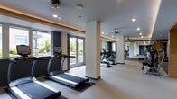 This contemporary fitness center offers a thoughtfully designed workout environment that balances functionality with modern aesthetics. The space is laid out on a single floor, featuring an open floor plan that allows for comfortable movement between equipment stations. Rows of cardio machines — including treadmills and ellipticals from True Fitness — are strategically positioned to take advantage of the large floor-to-ceiling windows, which flood the space with natural light and provide refreshing outdoor views of surrounding greenery. The neutral-toned flooring and light walls create a clean, energizing atmosphere, while the structural columns are finished to blend seamlessly with the overall design. Recessed lighting throughout the ceiling supplements the natural light, ensuring the space is well-lit at any time of day. Ceiling fans provide additional air circulation, contributing to a comfortable workout environment. Large mirrors line one wall, visually expanding the space and allowing users to monitor their form during exercise. The overall ambiance is sleek and professional, reflecting a commitment to providing a high-quality fitness experience within a well-maintained, modern facility.