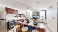 Located at 1004 N 10th St in Milwaukee, WI, this one-bedroom, one-bathroom unit offers an efficiently designed single-floor layout. The open-concept living and kitchen area serves as the heart of the space, featuring terrazzo flooring, a ceiling fan with light fixture, and multiple windows that bring in natural light. The kitchen is equipped with rich dark-wood cabinetry, granite-style countertops, stainless steel appliances including a full-size refrigerator and range, and open shelving for additional storage. A wood-topped kitchen island with built-in bar seating provides a functional dining and gathering area. The living space accommodates a media setup and benefits from bright, white walls that enhance the sense of openness. A short hallway connects the main living area to the private bedroom and bathroom. The bedroom features neutral carpet, clean white walls, and a ceiling fan, creating a calm and restful environment. The full bathroom is designed with accessibility in mind, incorporating grab bars near the toilet, a roll-in shower area, a dark-wood vanity with a white countertop, and a large mirror. Terrazzo flooring carries throughout the unit's common areas, tying the space together with a consistent, durable aesthetic.