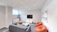 This single-floor space showcases a thoughtfully designed lounge area defined by a clean, modern aesthetic. The room features white walls and a smooth white ceiling fitted with multiple recessed spotlights, providing even, adjustable illumination throughout the space. A wall-mounted flat-screen television serves as a central focal point, positioned above a sleek white media console that complements the room's minimalist design language.

Seating is a highlight of the space, combining a generous grey sectional sofa with bold orange accent chairs and a matching ottoman, creating a dynamic contrast that adds energy and visual interest to the otherwise neutral palette. A low, white coffee table anchors the seating arrangement atop a grey area rug, reinforcing the contemporary style.

Natural light enters through a horizontal ribbon window along one wall, offering a subtle connection to the outdoors. Potted greenery adds a refreshing organic element, softening the clean lines of the interior. Along the opposite wall, a series of framed artwork is neatly arranged, contributing a gallery-like quality. White cabinetry provides practical storage while maintaining the space's cohesive, uncluttered appearance.
