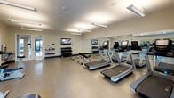 This fitness center is a thoughtfully designed, single-floor facility built to accommodate a comprehensive workout experience. The space is bright and open, featuring neutral-toned walls, smooth flooring, and overhead lighting that evenly illuminates the entire room. Large windows along one wall allow natural light to filter in, offering views of the outdoors and contributing to an energizing atmosphere.

The cardio section is well-appointed with multiple treadmills, elliptical trainers, and stationary bikes, each equipped with individual digital displays for an engaging workout. A dedicated strength training area features a multi-tiered dumbbell rack stocked with a full range of free weights, accompanied by an adjustable workout bench for versatile exercise options.

Floor-to-ceiling mirrors line key walls throughout the space, visually expanding the room and allowing users to monitor their form during exercise. Wall-mounted flat-screen televisions are positioned for easy viewing from various points in the room, adding an entertainment element to any workout session. A small shelf near the entrance provides space for personal items or informational materials. The overall design is clean, functional, and well-organized, making this fitness center a practical and inviting space for daily exercise.