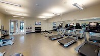 This fitness center is a thoughtfully designed, single-floor facility built to accommodate a comprehensive workout experience. The space is bright and open, featuring neutral-toned walls, smooth flooring, and overhead lighting that evenly illuminates the entire room. Large windows along one wall allow natural light to filter in, offering views of the outdoors and contributing to an energizing atmosphere.

The cardio section is well-appointed with multiple treadmills, elliptical trainers, and stationary bikes, each equipped with individual digital displays for an engaging workout. A dedicated strength training area features a multi-tiered dumbbell rack stocked with a full range of free weights, accompanied by an adjustable workout bench for versatile exercise options.

Floor-to-ceiling mirrors line key walls throughout the space, visually expanding the room and allowing users to monitor their form during exercise. Wall-mounted flat-screen televisions are positioned for easy viewing from various points in the room, adding an entertainment element to any workout session. A small shelf near the entrance provides space for personal items or informational materials. The overall design is clean, functional, and well-organized, making this fitness center a practical and inviting space for daily exercise.