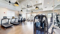 Located at 415 Curtis Pkwy SE in Calhoun, Georgia, this fitness center offers a thoughtfully designed exercise space that balances functionality with refined aesthetics. The room is defined by its striking coffered ceiling adorned with matte black pendant lights on gold fixtures and ceiling fans, lending a contemporary yet inviting atmosphere. Light wood-look flooring runs throughout, complementing the bright, neutral wall tones and contributing to a clean, energizing environment. Large windows along multiple walls flood the space with abundant natural light, while glass-paneled doors provide views toward the outdoor pool area, creating a seamless indoor-outdoor connection. A full wall of mirrors on one side visually expands the room and supports strength training activities. The fitness floor is comprehensively equipped with a row of Bodycraft treadmills, elliptical trainers, recumbent and upright bikes, multi-function cable and strength machines, and a variety of resistance and free-weight stations. A dedicated accessories area features stability balls and medicine balls for functional training. Wall-mounted flat-screen televisions are positioned for easy viewing from cardio equipment. The space is well-organized to accommodate a broad range of workout routines, making it a complete and polished fitness facility.